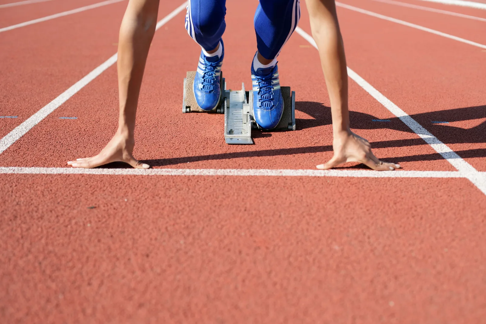 A sprinter crouched in the starting blocks on a red running track, hands on the line, seen head-on from track level.