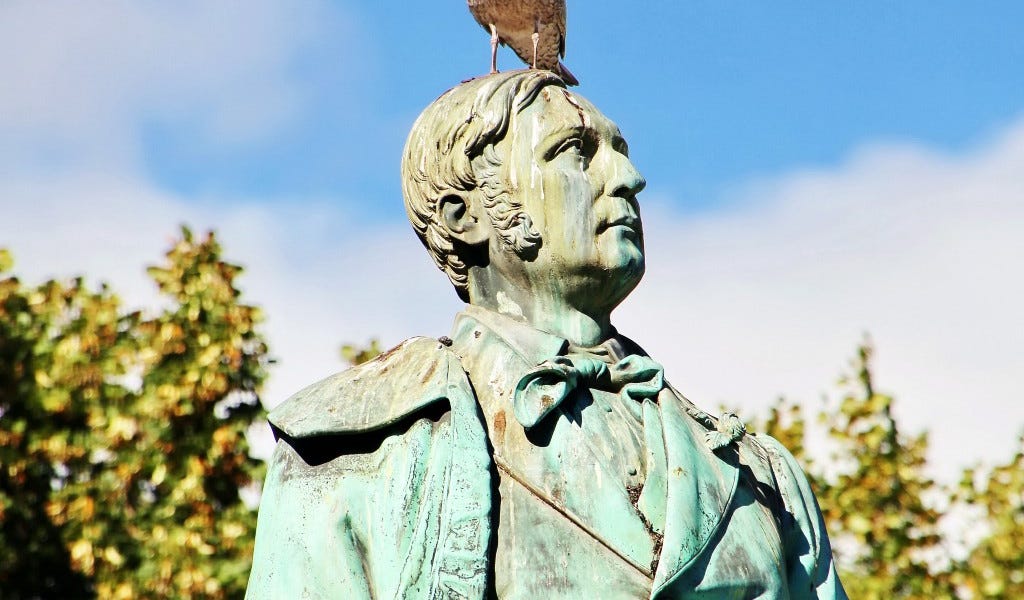 A bronze statue of a man in a bow tie, streaked with bird droppings, with a pigeon perched on his head.